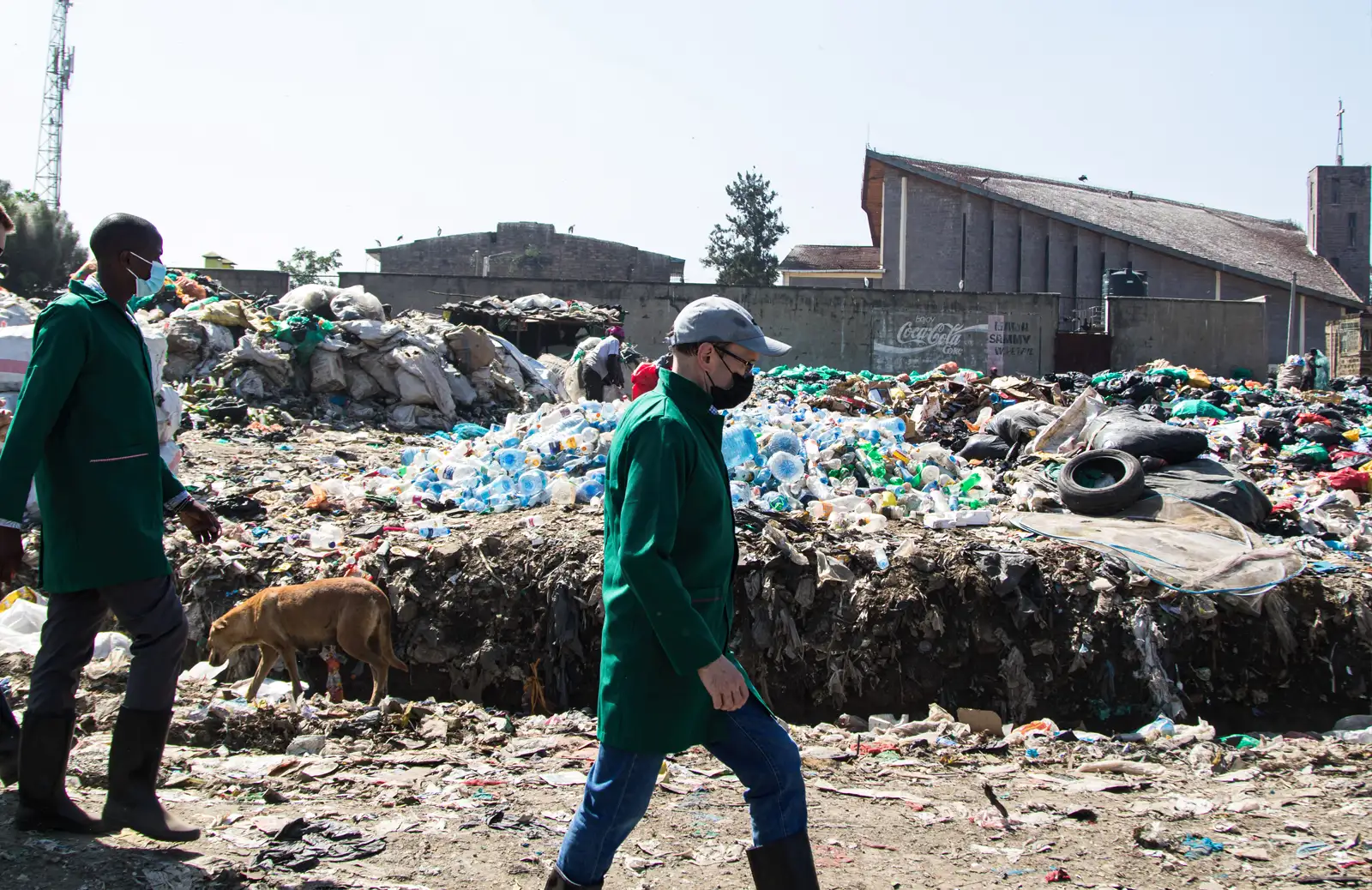 Two men walk through a dumping site that is filled with landfill, much of it plastics. They are wearing green protective coats and facemasks. In the background is a tan-coloured dog just making its way through the dump.