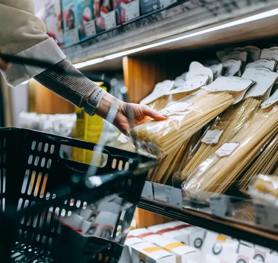 A woman‘s hand reaches for a packet of dry fettucine that is on a shelf in a supermarket aisle. In the foreground there is a black plastic shopping basket.