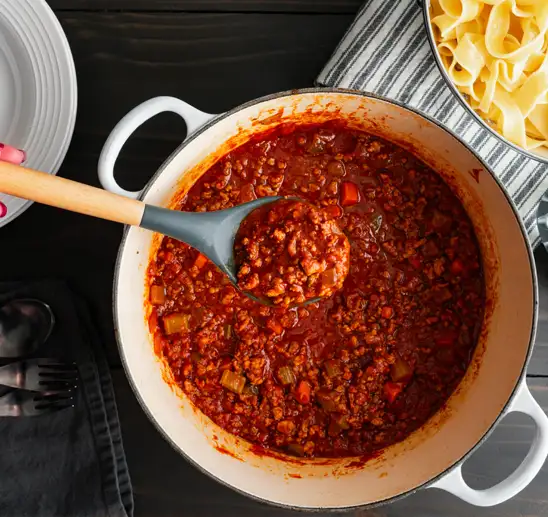A bird’s-eye view of a large pot of Bolognese sauce, with a pot of cooked pappardelle peeking in from the top left. A wooden-handled, plastic-headed ladle is lifting up a small amount of the sauce.