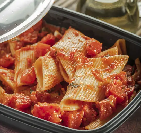 A close up of a dish of cooked manicotti pasta with a tomato-based sauce, in a black plastic container. A small amount of the clear plastic lid is tilted over the container.